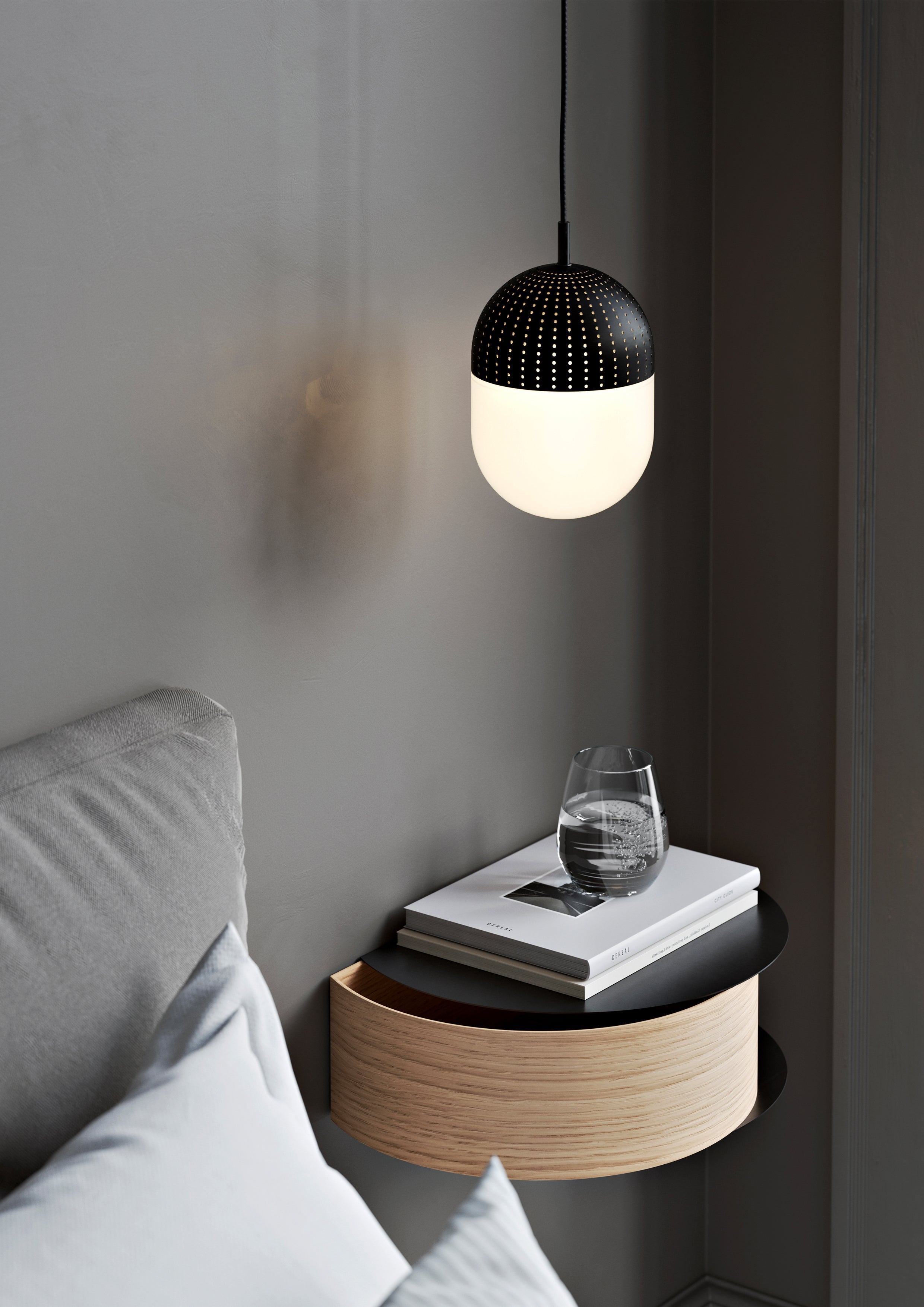 Pendant with perforated metal and opal glass shade hanging above wall-mounted oak shelf in bedroom beside bed.