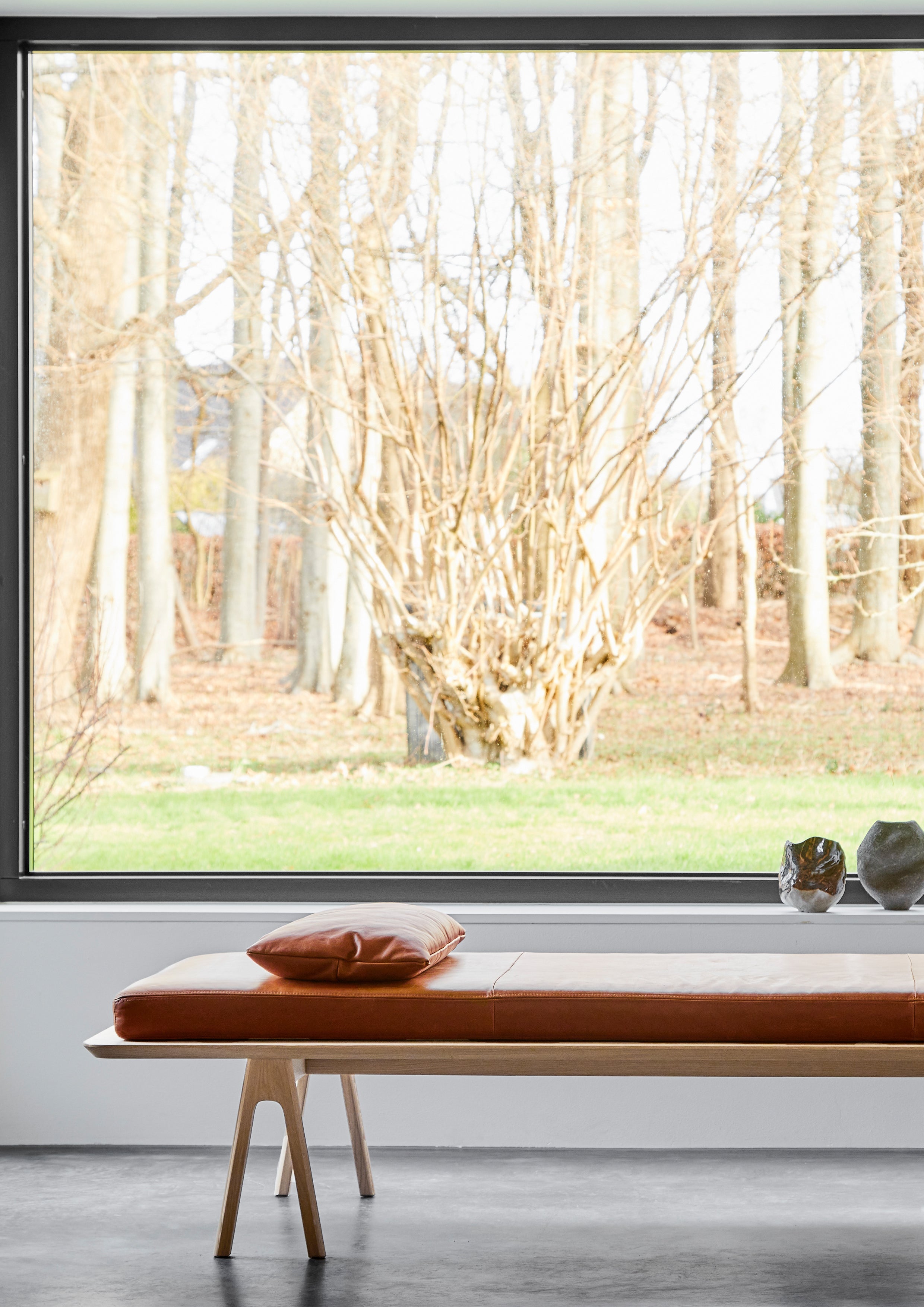 Daybed with white pigmented oak frame and cognac leather mattress in front of window with forest view.