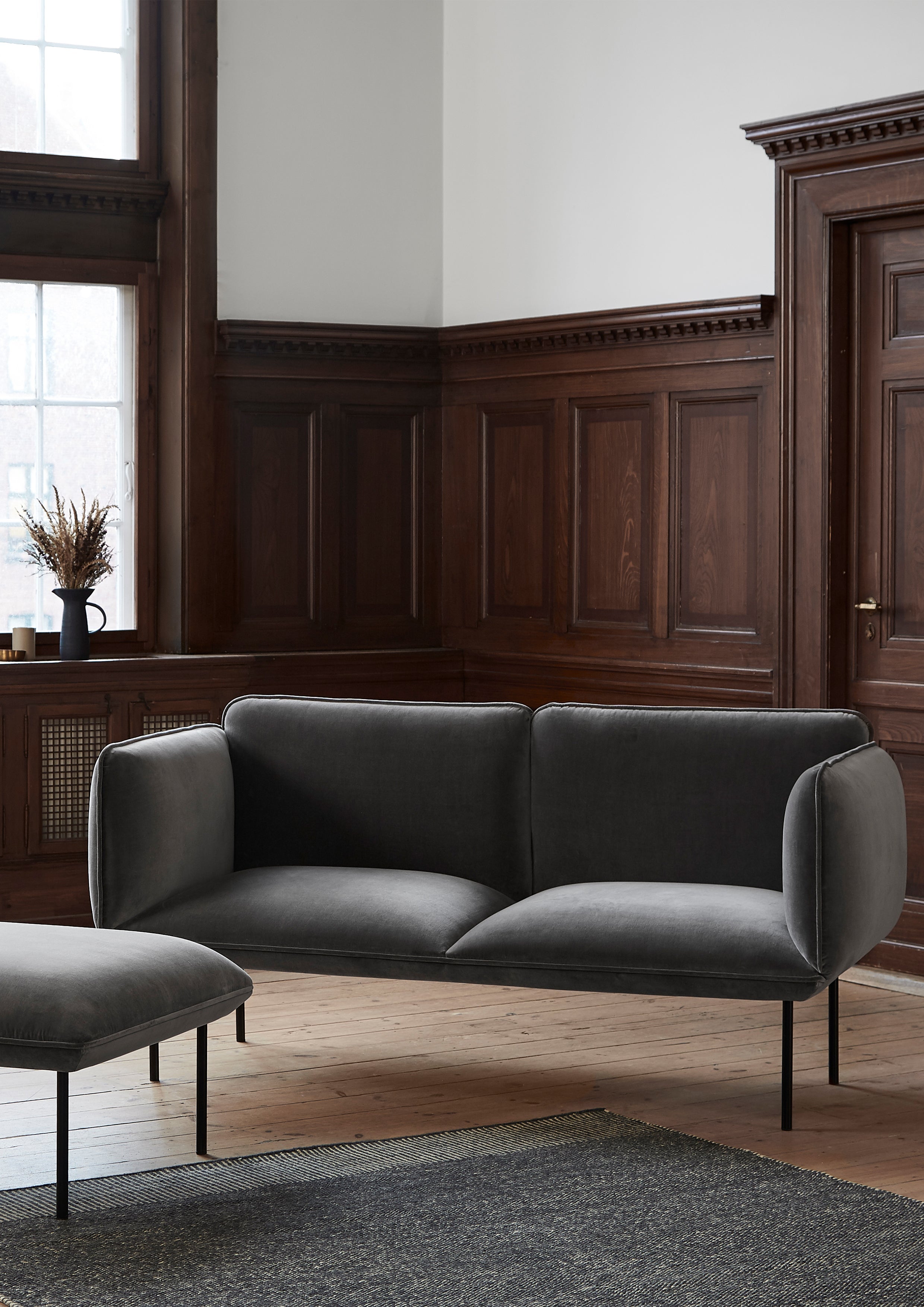 Grey velvet sofa in minimalist design placed in a room with tall dark wood wall paneling.