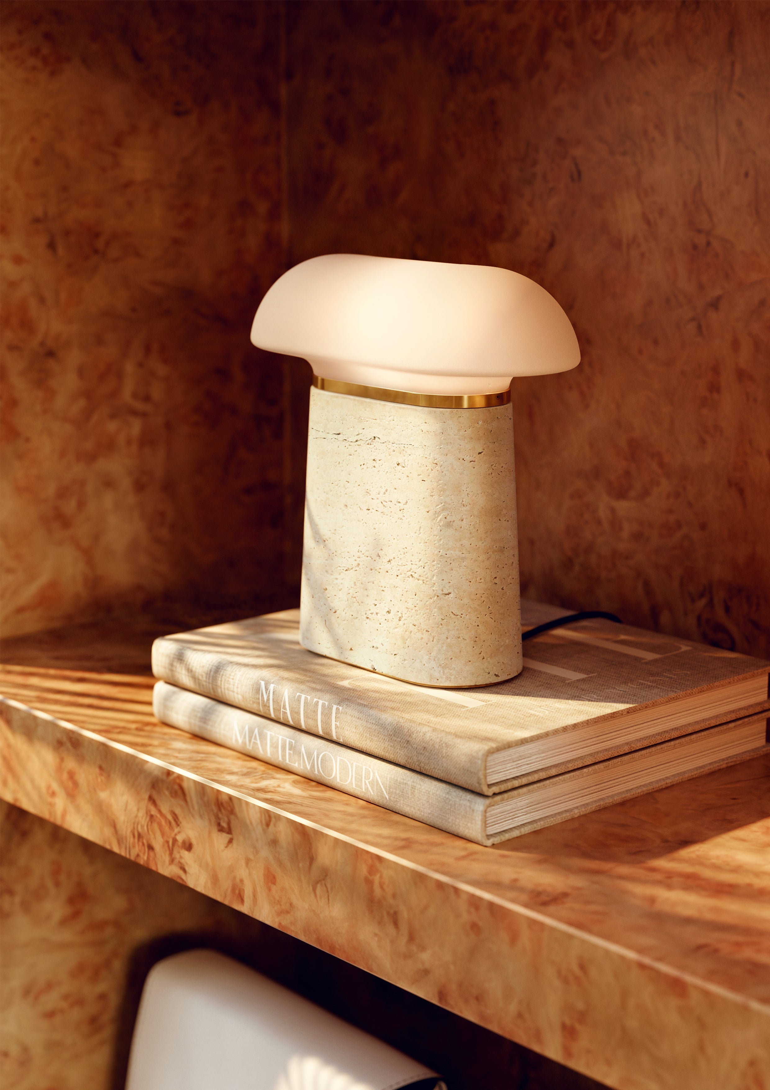 Table lamp with ivory travertine base, opal glass shade, and brass details, on books in shelving unit.